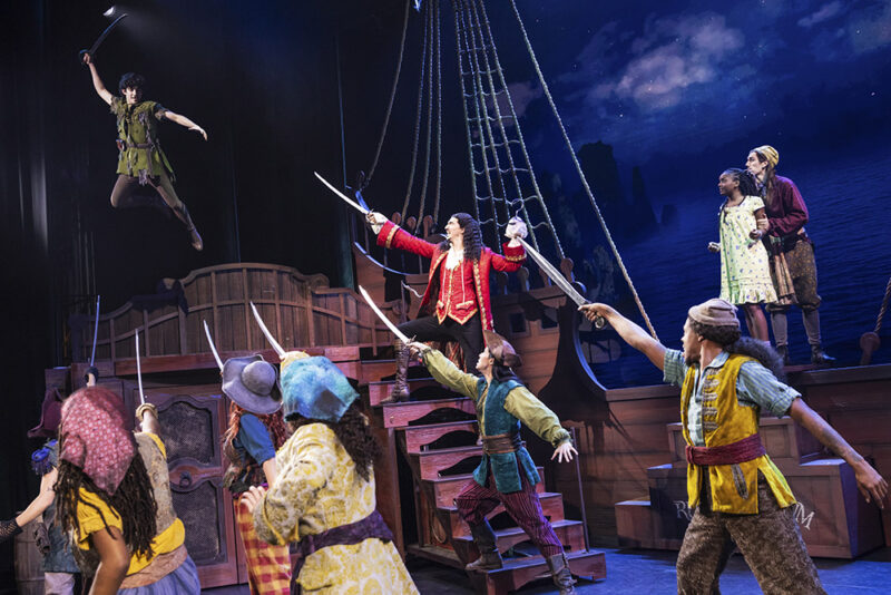 Nolan Almeida as Peter Pan, Cody Garcia as Captain Hook, Hawa Kamara as Wendy, and the cast of Peter Pan. Peter Pan flies above Captain Hook’s ship as Hook and his pirate crew all face him pointing their swords in his direction. Another pirate stands on the side of the ship holding Wendy in place.