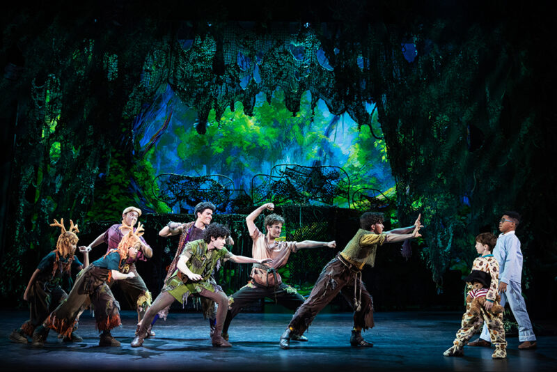 Nolan Almeida as Peter Pan and the cast of Peter Pan. Peter Pan and The Lost Boys crouch with their arms raised as they lean toward Michael and John who slightly lean away.