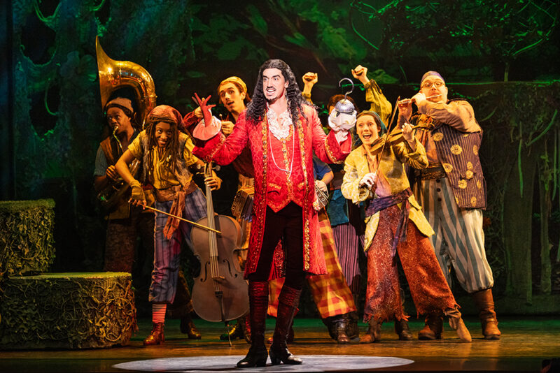 Cody Garcia as Captain Hook and the cast of Peter Pan. Hook stands center, both hand and hook up, singing as his group of swashbucklers plays instruments behind him.